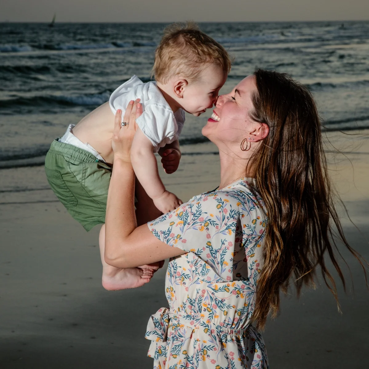 A mom holding up her baby, smiling happily, standing by the water's edge during a professional photo session on the beach in Fort Morgan, Alabama.
