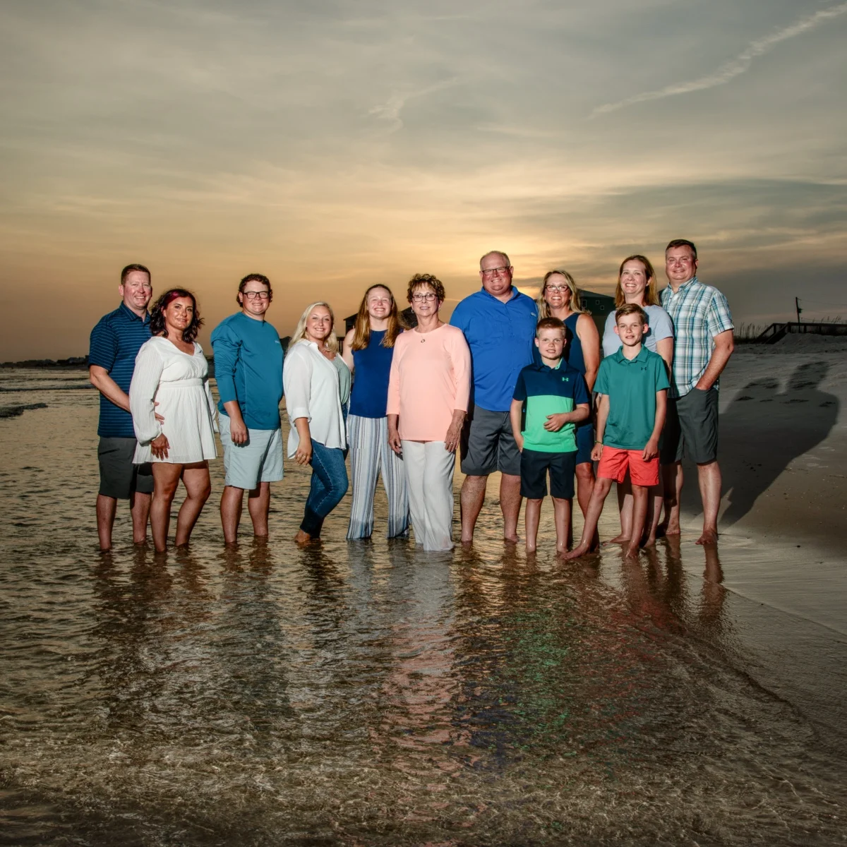 A sunset beach photo of a family of 12 standing in the water's edge during a photo session in Fort Morgan, Alabama.
