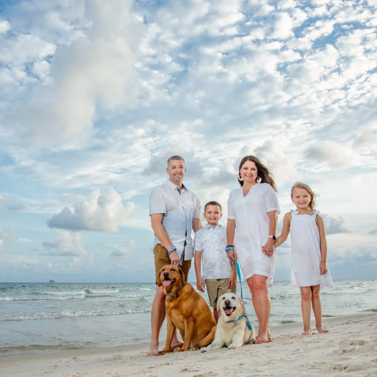 Family of four with their two dogs standing by the water's edge for a family photo session on the beach in Fort Morgan, Alabama.