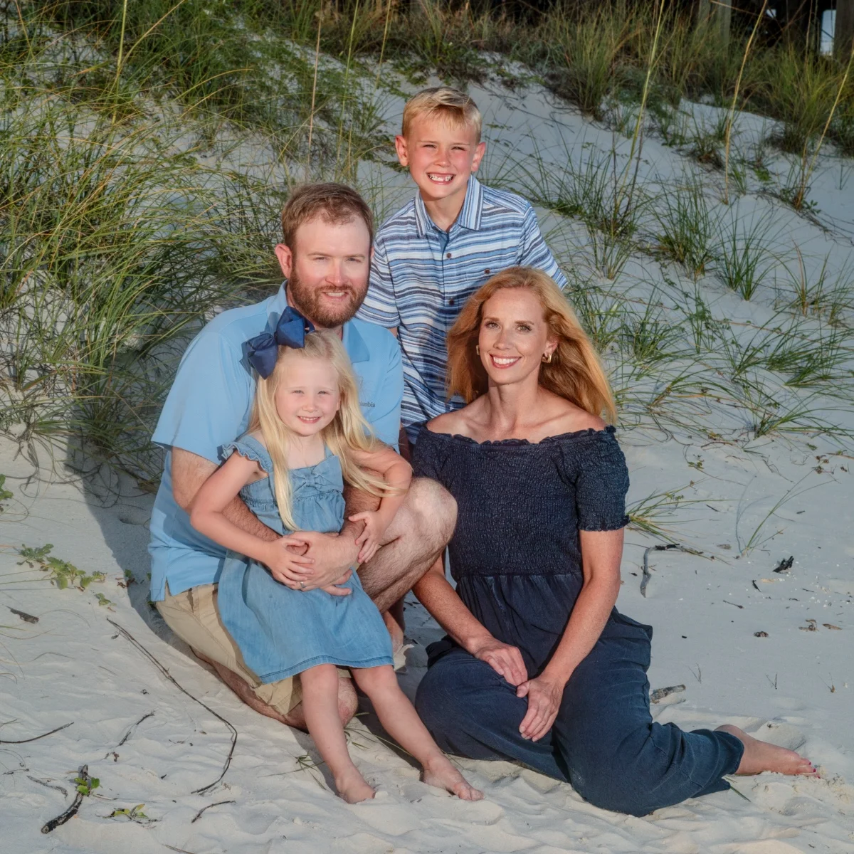 A family of five sitting in the sand dunes by the sea oats during a beach photo session in Fort Morgan, Alabama.