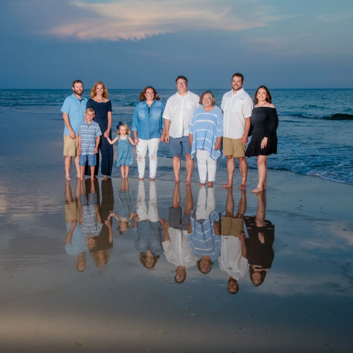 A family of nine standing by the water's edge with a beautiful reflection of them on the beach for a photo session in Fort Morgan, Alabama.