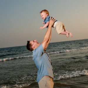 A dad holding his baby son up in the air at sunset by the water's edge in Fort Morgan, Alabama during a photo session.