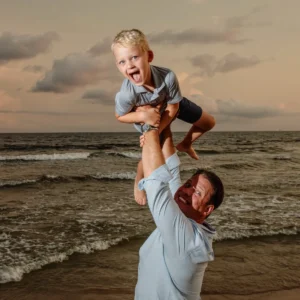 A dad holding his son up in the air, standing by the shoreline at sunset for a photo in Fort Morgan, Alabama.