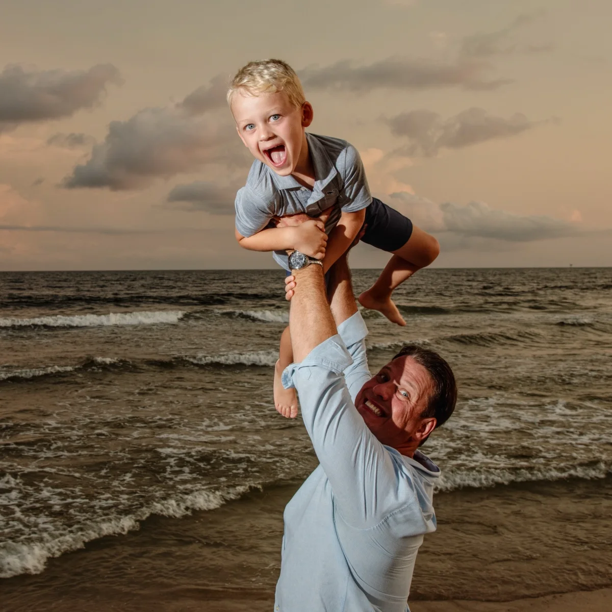 A dad holding his son up in the air, standing by the shoreline at sunset for a photo in Fort Morgan, Alabama.